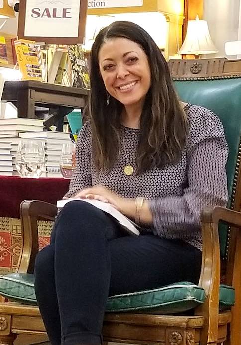 Stacy Dicker sitting in a chair in the Boulder Bookstore smiling with a book on her lap.