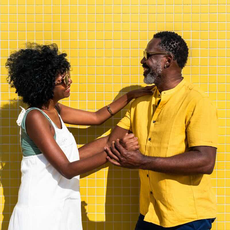 A mature black couple having fun while dancing in the sun next to a yellow brick wall.