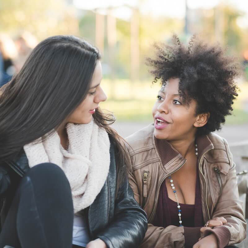 Two multiethnic women talking to each other outside in a park with light jackets on.